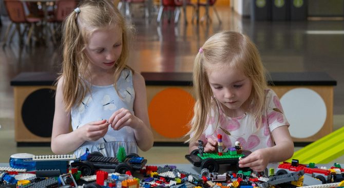 Two young children play with colorful building blocks on a table in a bright, spacious indoor play area.
