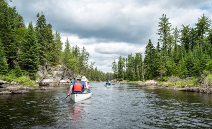 Two canoes travel down a river with tree-lined shores.