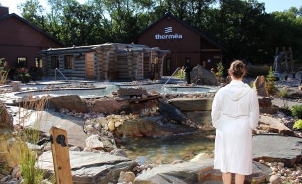 A lady in a white robe standing overlooking the outdoor pools and spa at Thermëa by Nordik Spa-Nature
